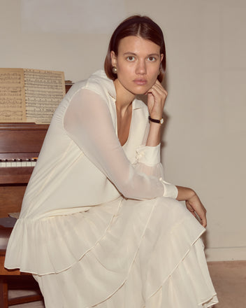 A woman wearing the Vendome Gown with French cuffs sits by a piano with sheet music, resting her chin on her hand.