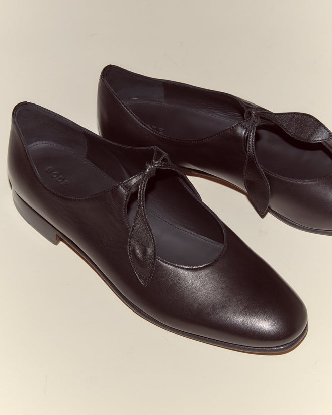 The Verbena Shoes - Black are black leather shoes with a front tie detail, inspired by classic Mary Janes, shown on a light beige surface.