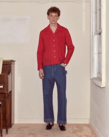 Indoors near a piano and window, a person wears the Voile Long Sleeve Shirt - Red, styled with blue wide-leg jeans and black shoes.