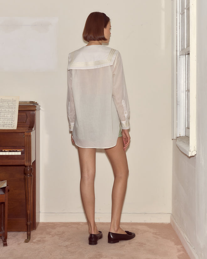 A woman stands indoors facing a wall, wearing the Voile Marina Shirt with dark loafers near a piano and window.