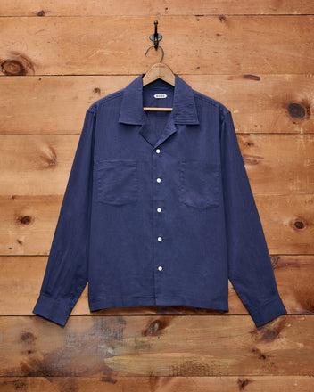 A Voile Long Sleeve Shirt in navy, featuring a boxy silhouette, hangs on a black hook against a wooden wall.