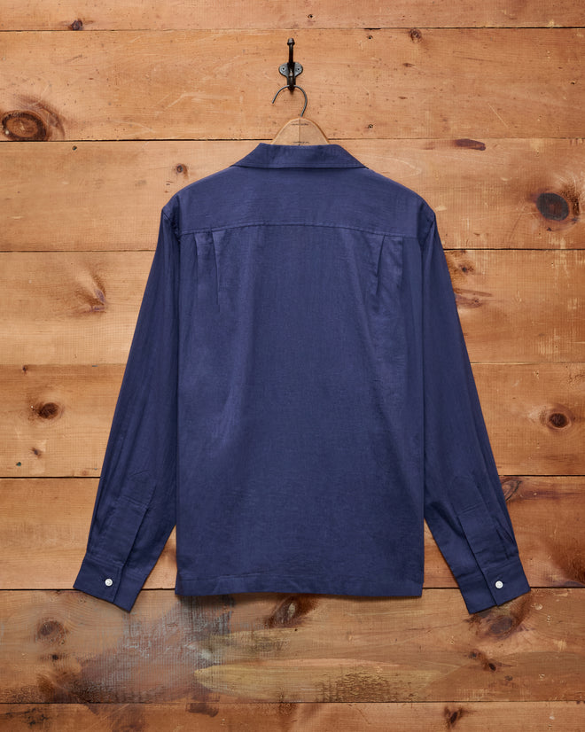 The Voile Long Sleeve Shirt - Navy, featuring a boxy silhouette and crafted from cotton, is displayed from the back hanging on a wall hook against a wooden backdrop.