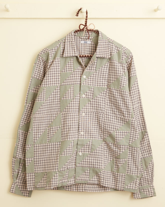 A Wandering Lover Quilt Long Sleeve Shirt - Mint, featuring a patchwork design in shades of green and beige that evokes the charm of an antique quilt, hangs on a peg against a cream-colored wall.