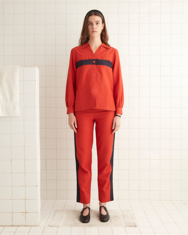 A person wearing the Waterfront Shirt, designed with a red pattern and black stripes reminiscent of a 1940s women's blouse, stands in a tiled room facing forward.