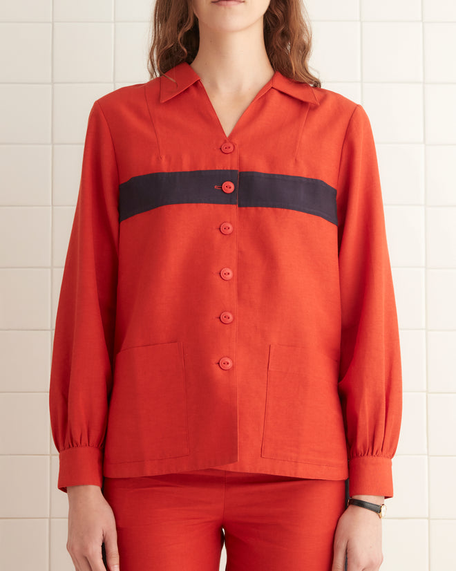 A person wearing the Waterfront Shirt, designed with a red pattern and black stripes reminiscent of a 1940s women's blouse, stands in a tiled room facing forward.