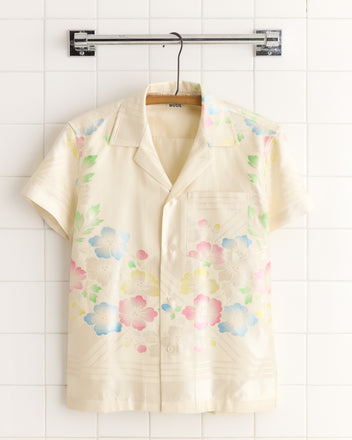 The Waterlily Stencil Shirt - XS, a short-sleeve top with a delicate cream pastel floral print, hangs on a hanger against a white tiled wall.