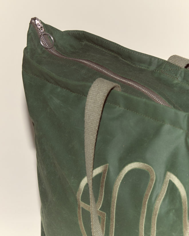 The Waxed Cotton Zip Tote - Green features light handles, a zip closure, and an embroidered Bode logo. Crafted in Italy from green waxed cotton, it's shown against a plain background.