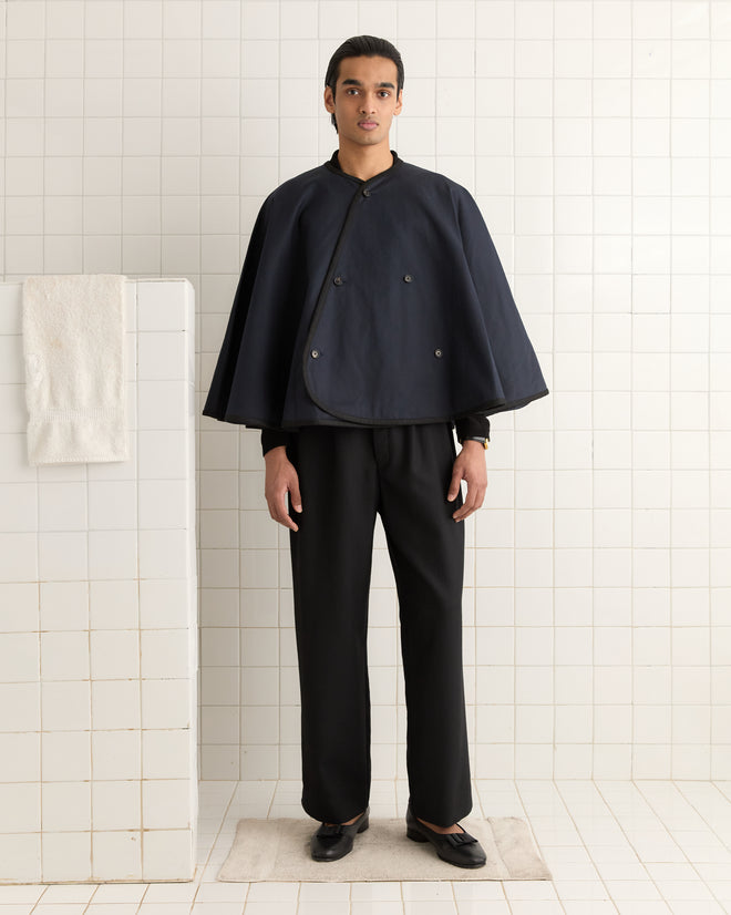 In a tiled room, someone wears the Waxed Herringbone Rain Cape in navy with matching pants, beside a towel on a wall hook.