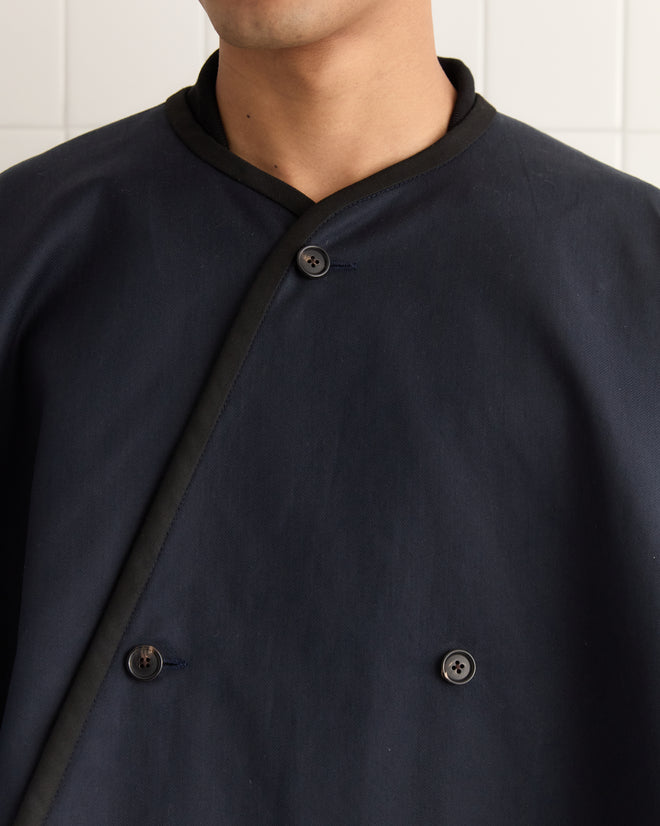 A person wearing a navy Waxed Herringbone Rain Cape, featuring an asymmetrical button design, stands against a tiled wall.