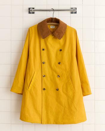 The Waxed Peacoat - Yellow, featuring a brown collar and crafted from waxed cotton, hangs stylishly on a tiled wall.