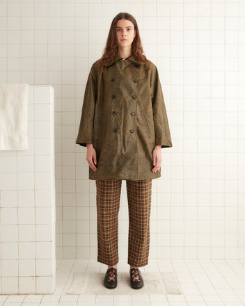 A person with long hair wearing a brown Waxed Swing Peacoat and plaid pants stands on a white tiled floor, with a white towel hanging on a rack in the background.