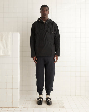 A person standing in a tiled room, wearing a sleek black Waxed Tie Anorak, dark pants, and black shoes with socks.