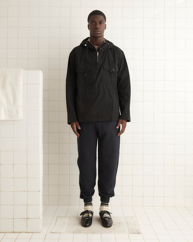 A person standing in a tiled room, wearing a sleek black Waxed Tie Anorak, dark pants, and black shoes with socks.