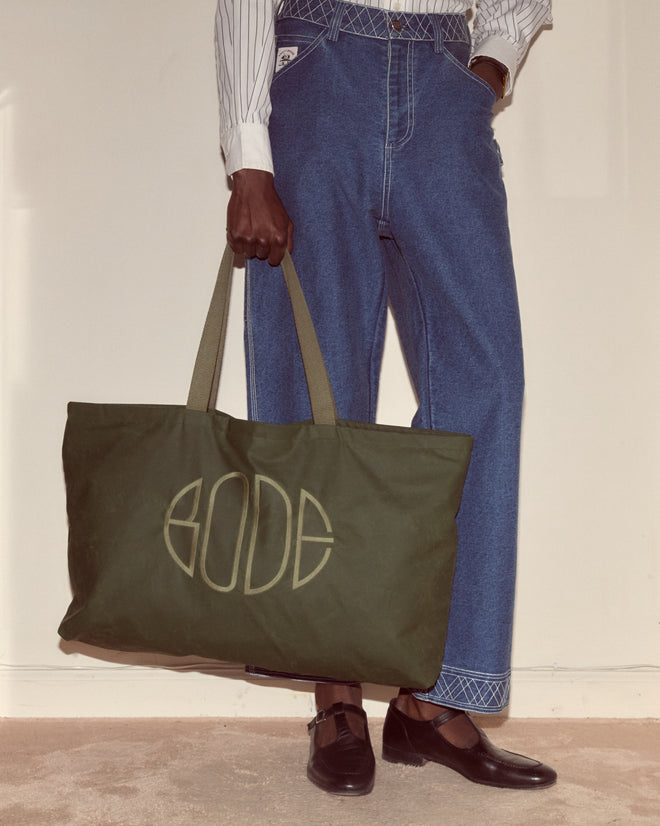 A person in blue jeans holds the Waxed Cotton Zip Tote - Green with olive handles and an embroidered Bode logo, standing on a beige carpet.