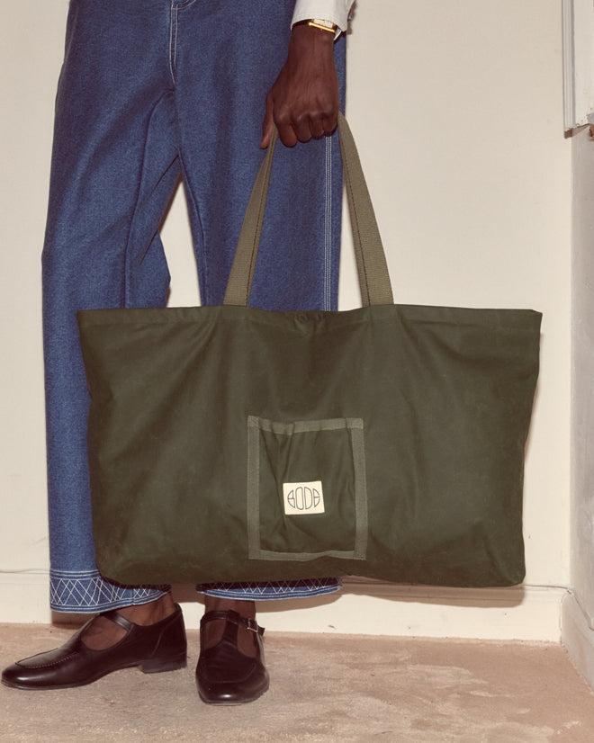 A person in blue jeans holds the Waxed Cotton Zip Tote - Green, featuring a large size, front pocket, embroidered Bode logo, and paired with black shoes.
