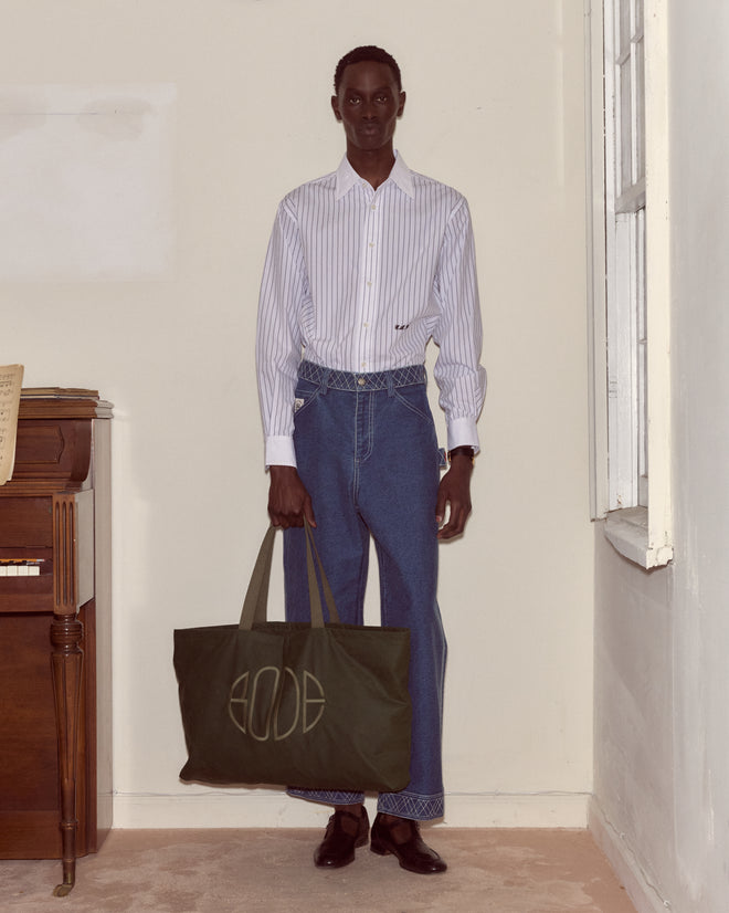 A man in a striped shirt, blue jeans, and black shoes holds the Waxed Cotton Zip Tote - Green indoors.