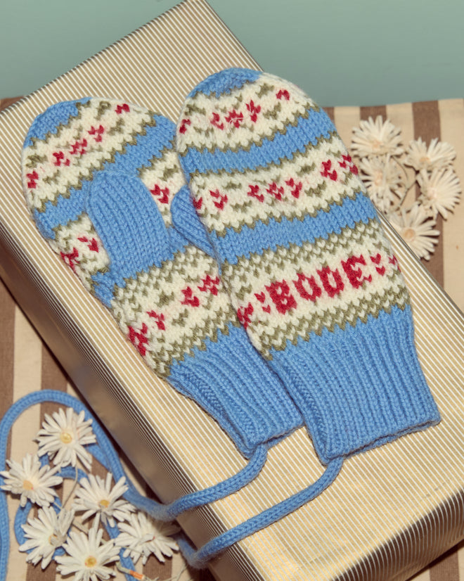 Wedgewood Mittens, crafted from virgin wool with blue, white, red, and green patterns, are displayed on a striped gift box decorated with white flowers.