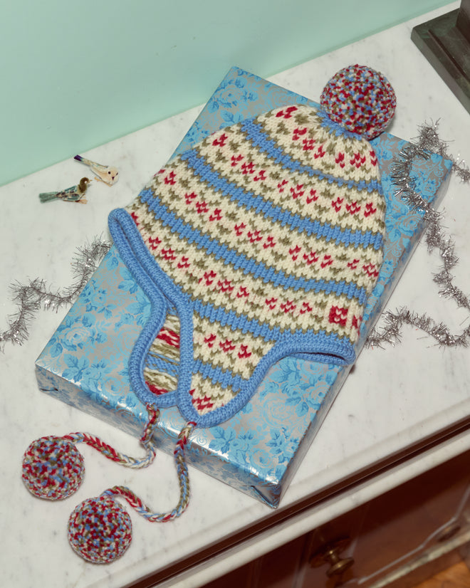 The Wedgewood Pom Hat, crafted from virgin wool and featuring blue, cream, and red stripes with pom-poms, is displayed on a blue gift box.