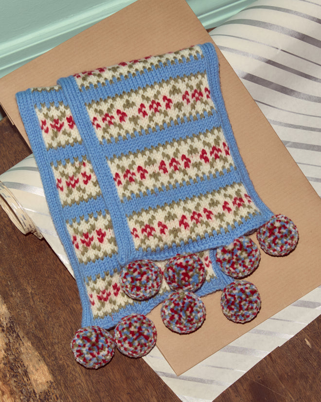 The Wedgewood Scarf, a vintage knit with blue edging and circular pom-poms, is displayed on brown paper and a wood floor.