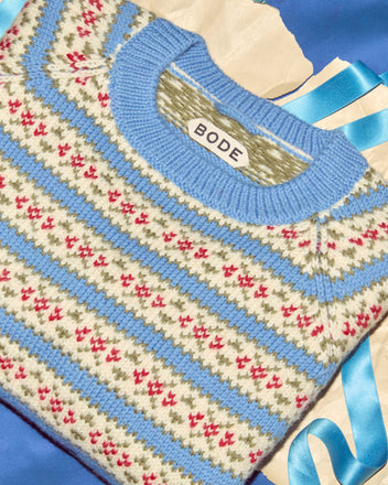 The Wedgewood Sweater is a folded BODE virgin wool knit featuring blue, white, red, and green patterns on blue fabric with a ribbon—ideal for fans of distinctive vintage-inspired style.