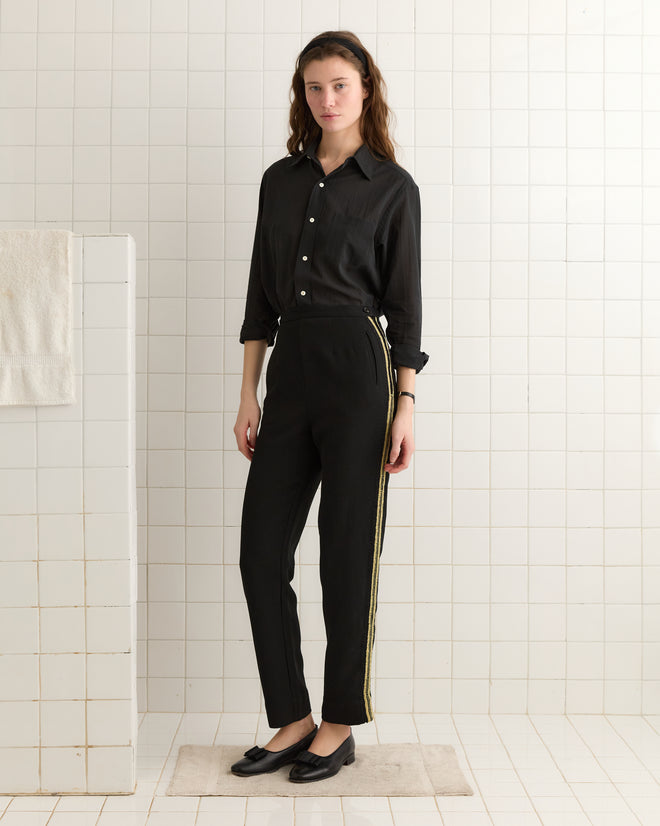 A person wearing Weedy Crochet Trousers in black with gold stripes and a straight leg design is standing in a tiled room with a white towel hanging nearby.