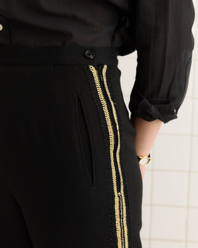A person stands against a tiled wall, wearing Weedy Crochet Trousers featuring gold zipper details and a black shirt.