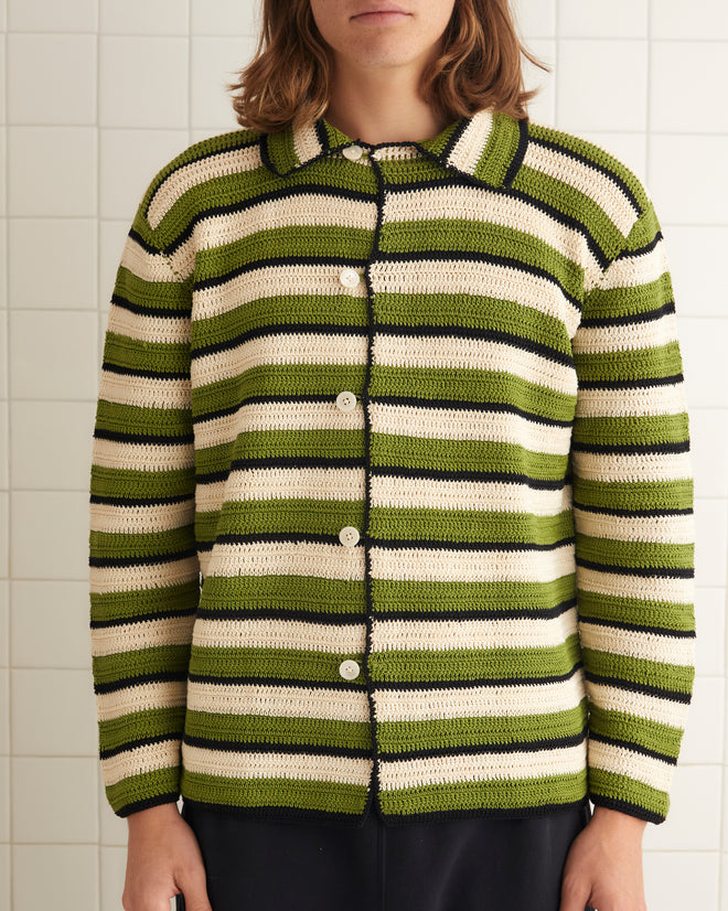 A person is standing in a tiled room wearing a Weedy Crochet Long Sleeve Shirt with green and white stripes that resembles a 1920s tunic, paired with black pants and black shoes. A white towel hangs on the nearby wall.