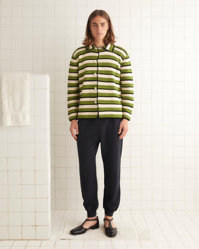 A person is standing in a tiled room wearing a Weedy Crochet Long Sleeve Shirt with green and white stripes that resembles a 1920s tunic, paired with black pants and black shoes. A white towel hangs on the nearby wall.