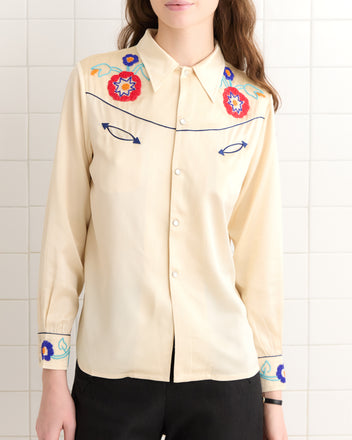 A woman wearing the Garden Party Western Shirt, featuring cream-colored fabric, floral embroidery, and pearl snap buttons, stands against a tiled wall.