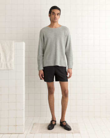 A person stands in a white-tiled room, draped in an elegant Wiley Mathers Weymouth Sweater of gray knitwear, paired with black shorts and shoes.