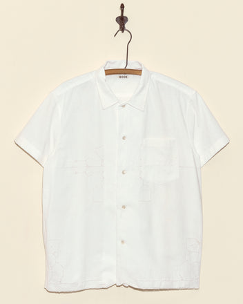 A White Hydrangea Short Sleeve Shirt (size L) with a chest pocket hangs on a hook against a beige wall, highlighting its vintage charm and unique drawnwork details.