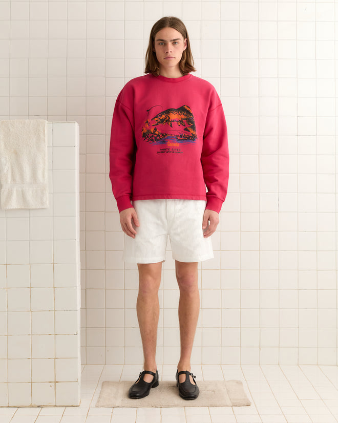 In a tiled bathroom, someone with long hair models the red White River Crewneck along with white shorts and black shoes.