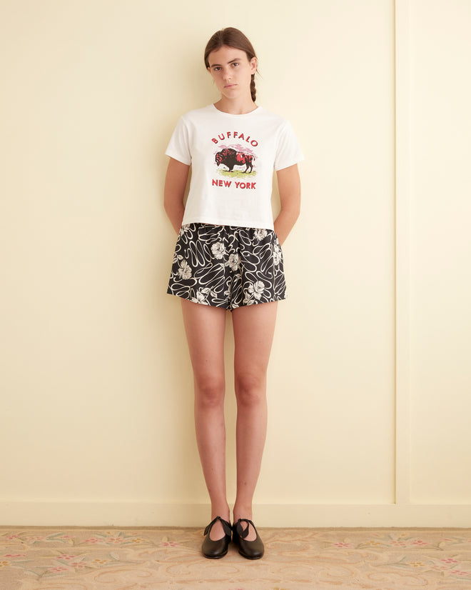 A person stands against a cream wall wearing a white t-shirt and White Rose Shorts.