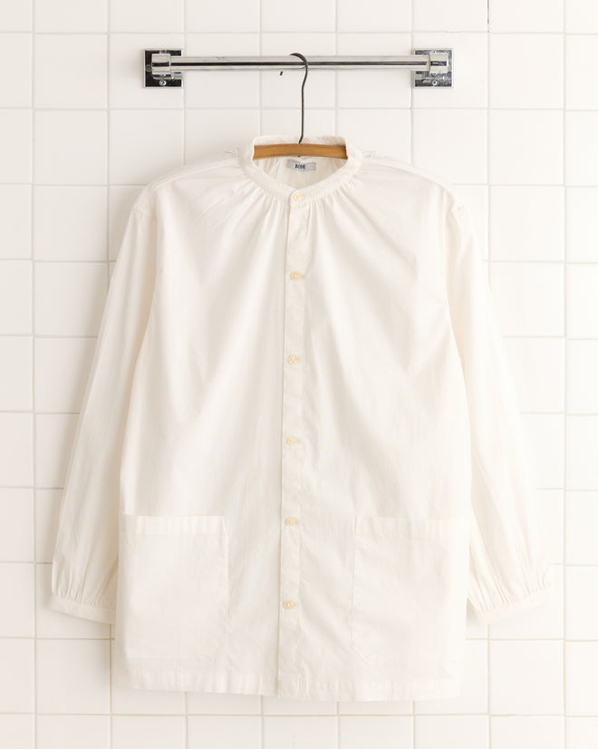 The White Smock Shirt, crafted from 100% cotton, is a long-sleeve button-up resembling a workmen's smock, hanging on a hanger against a tiled wall.