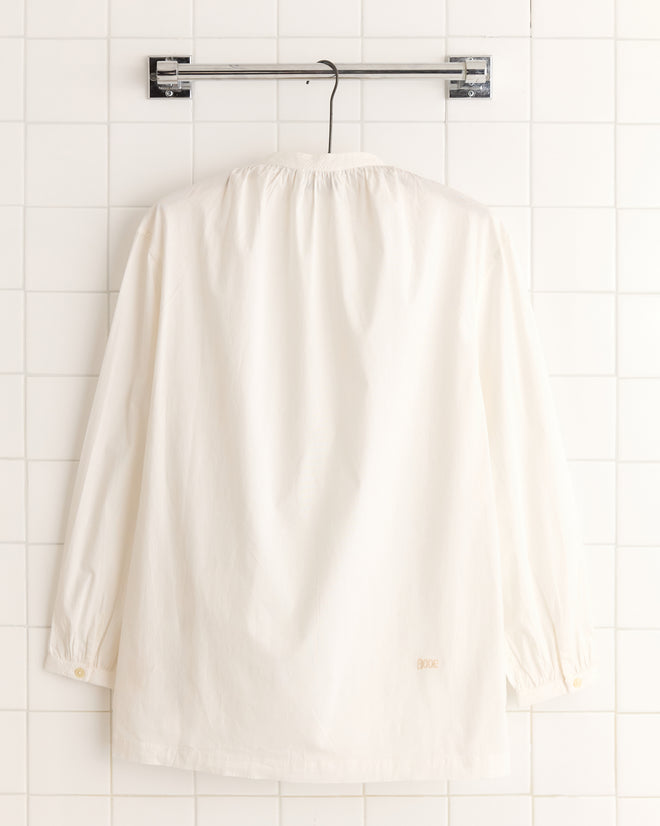 A White Smock Shirt, crafted from 100% cotton, hangs on a metal hanger against a tiled wall.