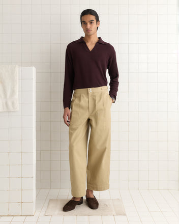 A person stands on a white tiled floor in a room with tiled walls, wearing a dark long-sleeve shirt and Wide Leg Snap Trousers - Khaki, reminiscent of vintage Australian military pants with side buckle waist adjusters.