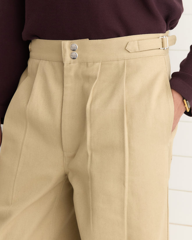 A person is wearing the Wide Leg Snap Trousers in Khaki with pleats and side buckle waist adjusters, paired with a dark sweater, standing with hands in pockets against a white background.