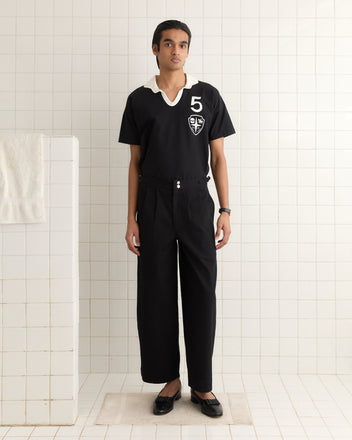 A person in black Wide Leg Snap Trousers stands in a tiled room with white walls, while a towel hangs nearby on a rail.