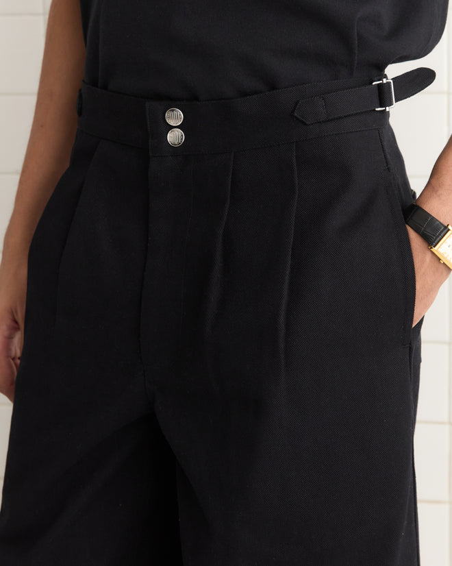 Sporting the Wide Leg Snap Trousers - Black with side buckle waist adjusters that echo Australian military style, the individual pairs them with a gold wristwatch elegantly gracing their left wrist.