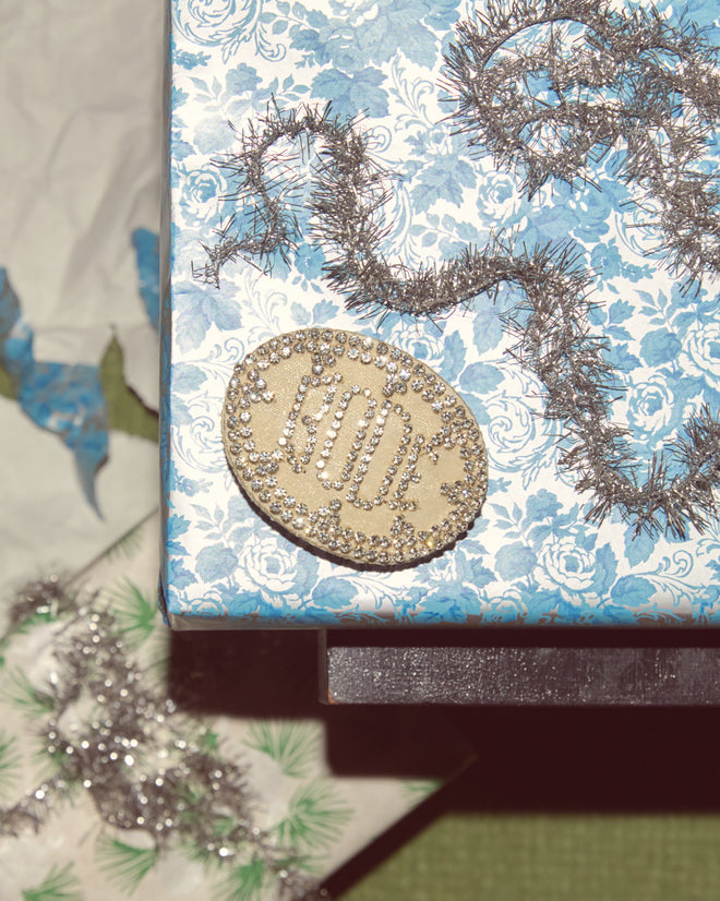 A gold and silver “NOEL” pin on a blue floral gift box with silver tinsel, paired with the Willow Barrette - White for a festive touch.