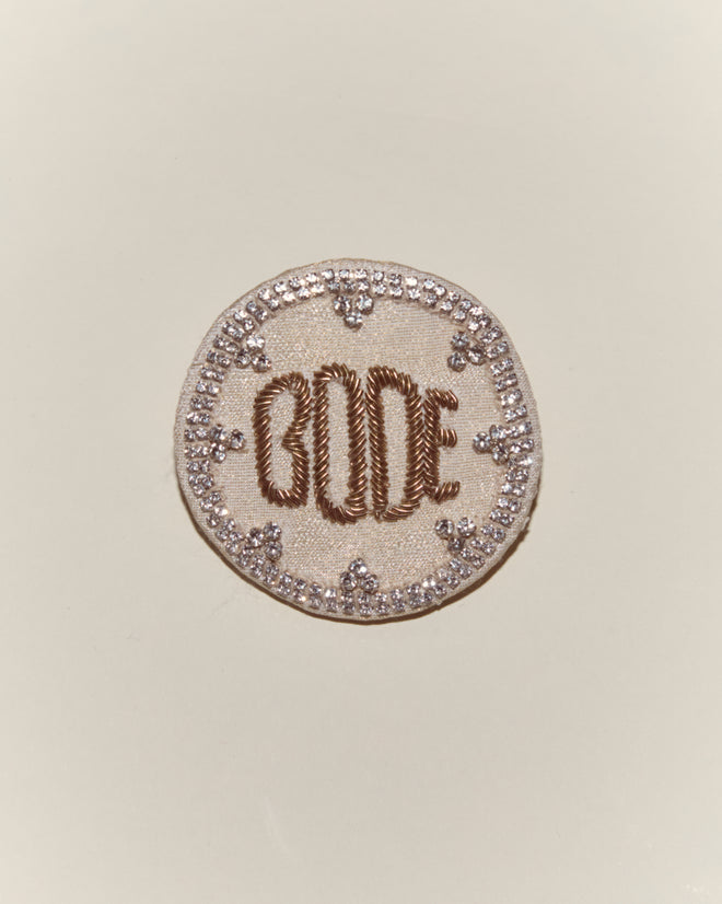 The Willow Brooch - White is a round embroidered accessory inspired by Bode, featuring "GOOD" in brown thread and a border of sparkling silver sequins.