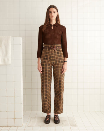 A person stands in front of a white tiled wall, wearing a dark red 1920s crocheted top with a keyhole neckline and plaid high-waisted pants. They are paired with black shoes featuring red laces. A towel hangs on the left.