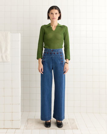 A person in a 1940s style Willows Pullover - Green and blue jeans stands in a tiled room, their vintage look accentuated by a keyhole button on the back.