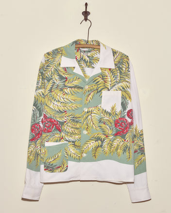 The Windy Fronds Shirt - S/M, featuring unique green, yellow, and red leaf patterns, hangs on a hook against a cream wall.