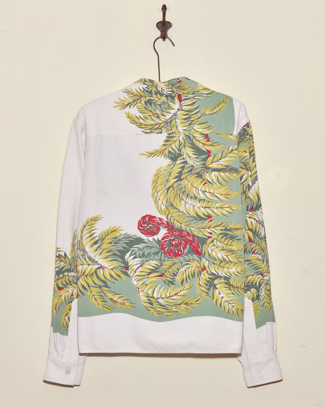 The Windy Fronds Shirt - S/M is a white vintage tablecloth shirt with green, yellow, and red floral print, displayed hanging on a wall hook from the back.