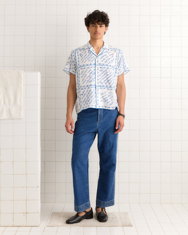 A man stands on a mat in a tiled room, wearing the Wine Poems Short Sleeve Shirt, blue jeans, and black shoes.
