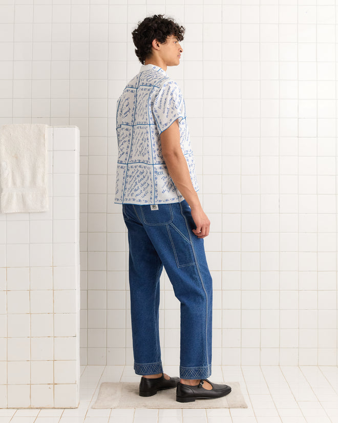 A person in blue jeans, the Wine Poems Short Sleeve Shirt, and black shoes stands in a tiled room.