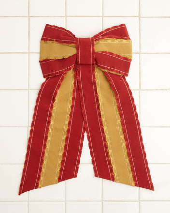 The Winner's Sash Bevel Bow, adorned with red and gold ribbons, elegantly hangs on a tiled wall, adding a touch of historical reproduction charm.