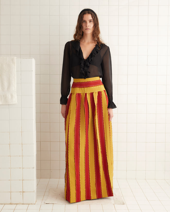 In a tiled room, a woman sports a black sheer blouse paired with the striking Winner's Sash Skirt in red and yellow stripes, accentuated by a delicate Victorian bow at the collar.