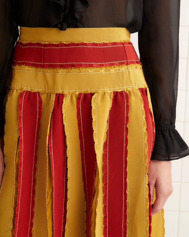 In a tiled room, a woman sports a black sheer blouse paired with the striking Winner's Sash Skirt in red and yellow stripes, accentuated by a delicate Victorian bow at the collar.